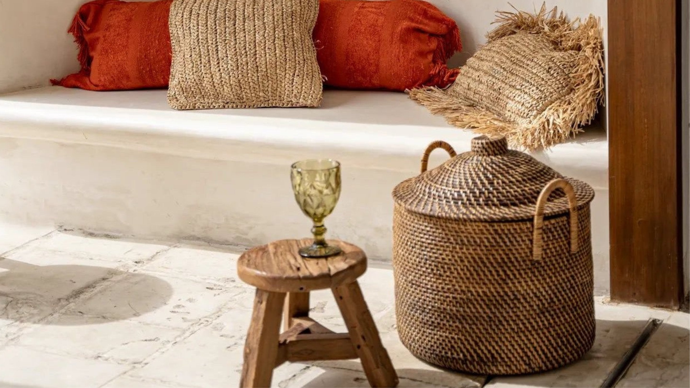 Bilden visar en träpall med ett grönt glas bredvid en stor flätad korg. I bakgrunden finns en sittplats med naturfärgade och orange kuddar på ett ljust underlag.