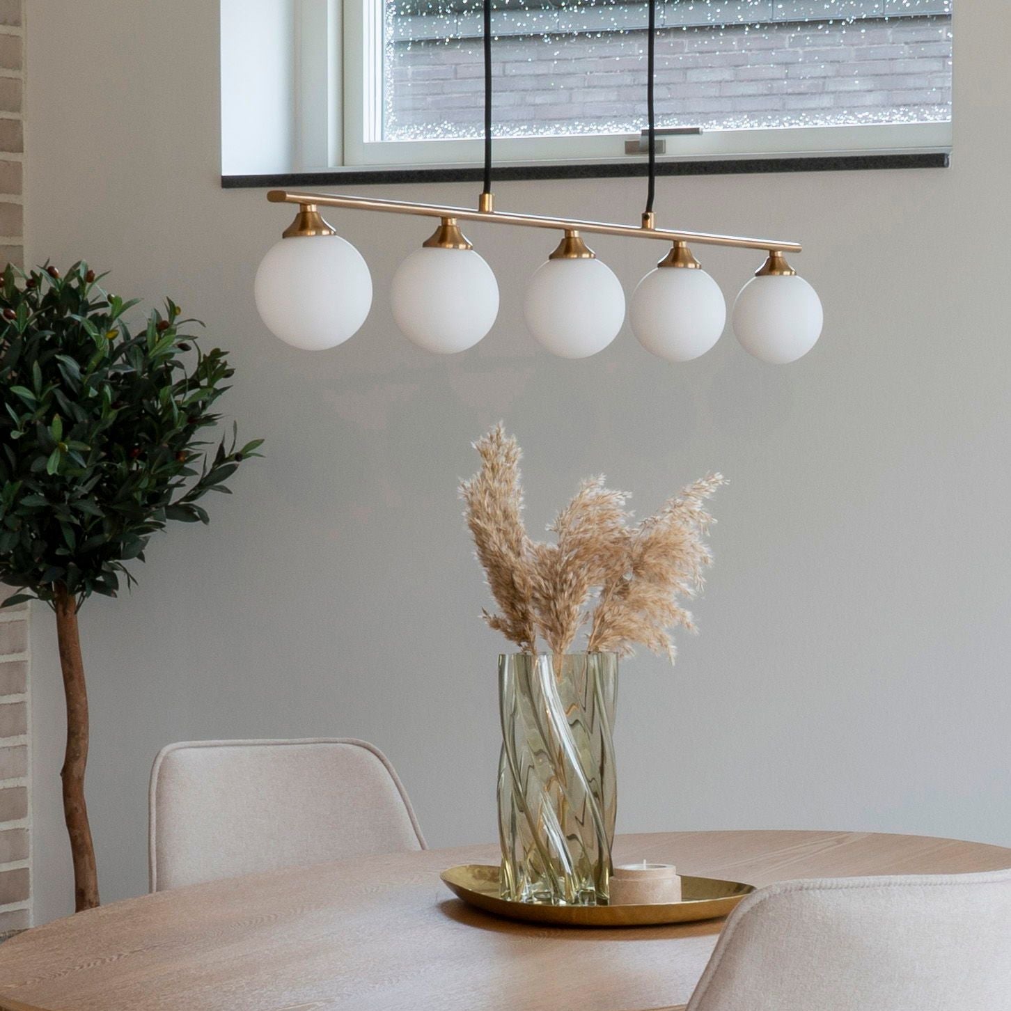En modern matplats visar upp ett runt träbord med en glasvas med torkat pampasgräs på en guldbricka. Ovanför den hänger House Nordics Halifax Pendellampa i Mässing med Glasglober. En krukväxt nära fönstret ger en touch av natur.