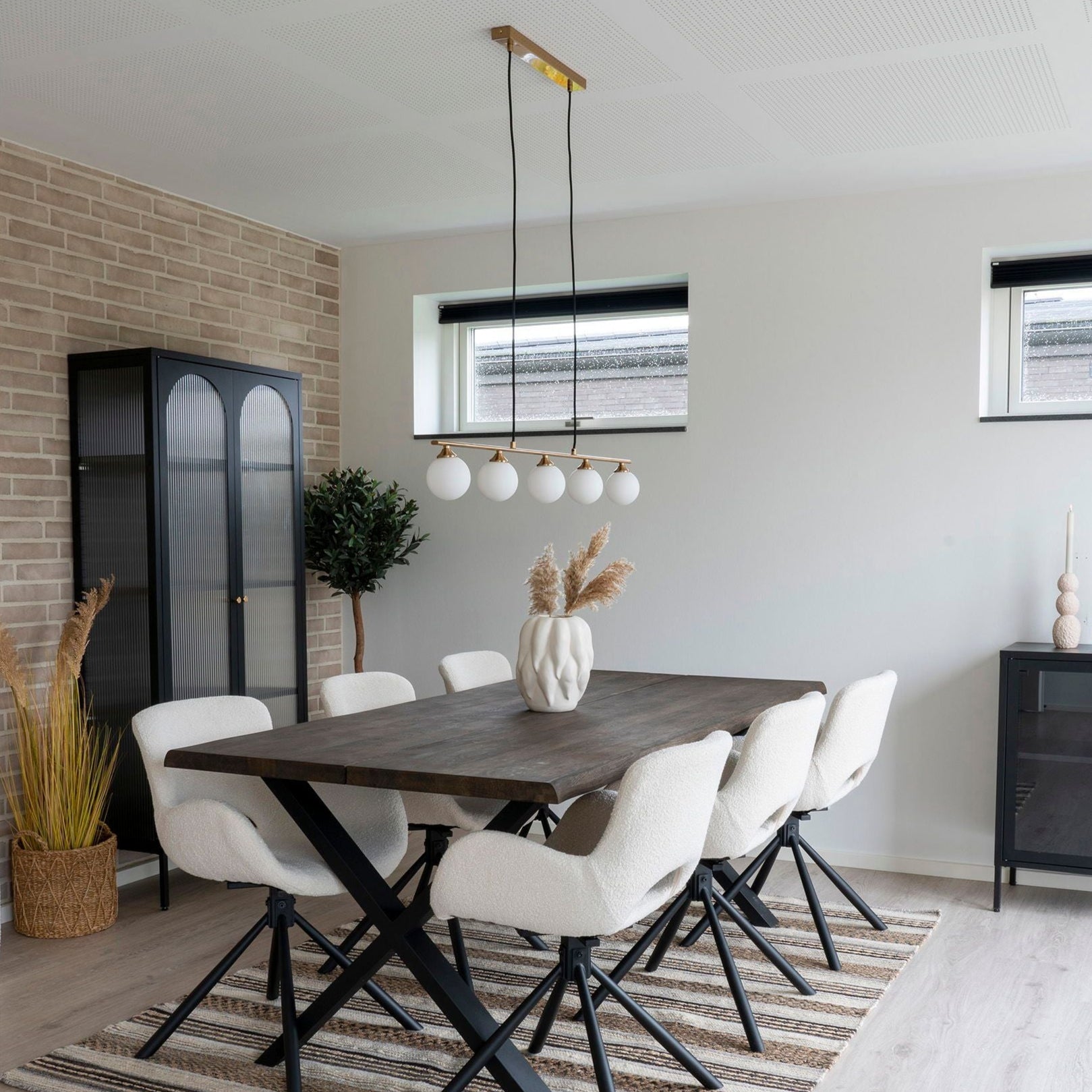En modern matsal har ett mörkt träbord med sex vita vadderade stolar på en randig matta. The House Nordics Halifax Pendellampa i Mässing hänger elegant ovanför och kastar ett varmt ljus. Ett svart skåp och krukväxt sitter mot tegelväggen, kompletterat med dekorativa torkade blommor.