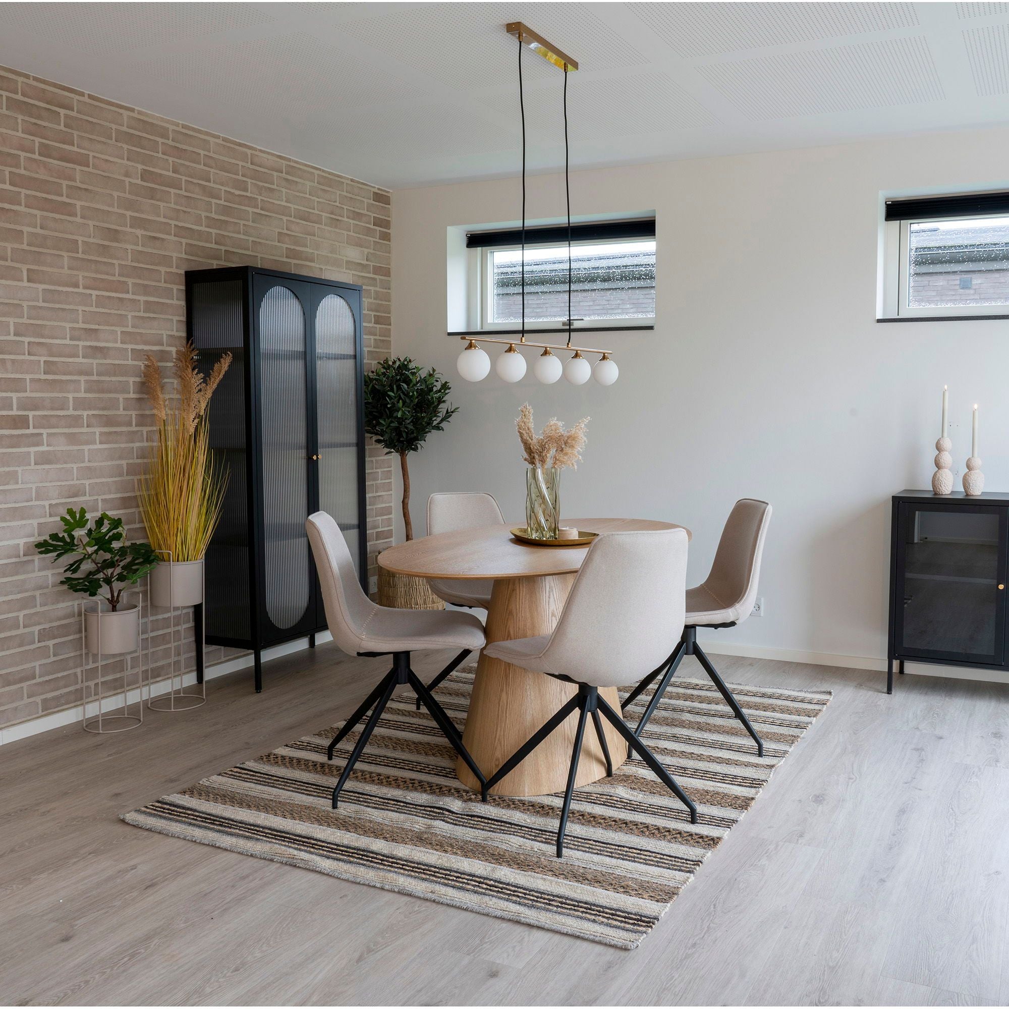 En modern matsal har ett runt träbord med fyra beiga stolar på en randig matta. En Halifax Pendellampa av House Nordic, i mässing med glasklot, hänger ovanför. Den kompletterar tegelväggen, det svarta skåpet, växterna och de stora fönstren för att skapa en mysig atmosfär i neutrala toner.