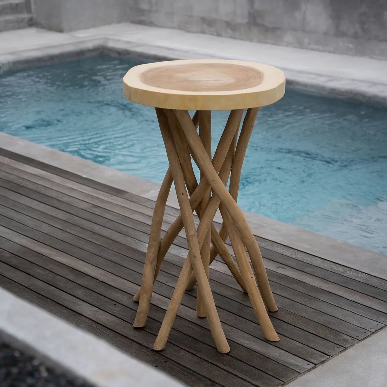 Gili barbord i teak från Bazar Bizar, med en naturlig finish och sammanflätade grenliknande ben, står på ett däck bredvid en pool. Poolens blå kontrast mot bordets jordnära toner, skapar en atmosfär av tropisk lyx och ö-vibbar.