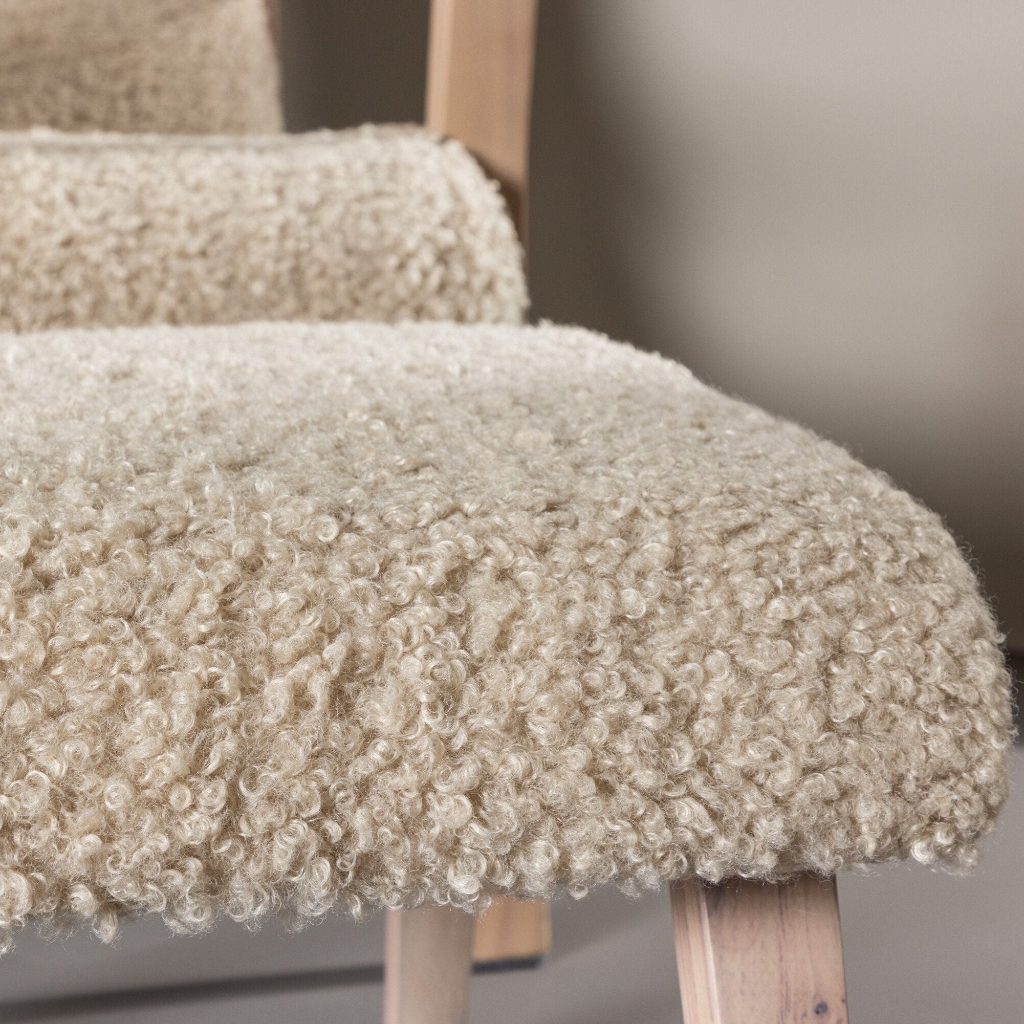 En närbild av Fårö fåtölj av Venture Home visar dess beige pälsliknande polyestertyg och ljusa träben som förstärker dess nordiska elegans. Skandinavisk-designen kombineras med plyschtyg för en mysig känsla, medan den suddiga bakgrunden ger djup till dess struktur och detaljer.