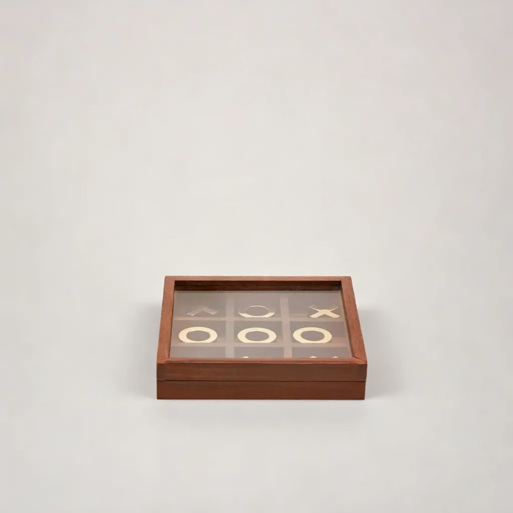 Tre i Rad Spel i Träask - Tic Tac Toe av Margit Brandt är ett spelset i trä med ett glaslock som visar tre O i rad bland spridda X på ett ljusgrått rutnät.