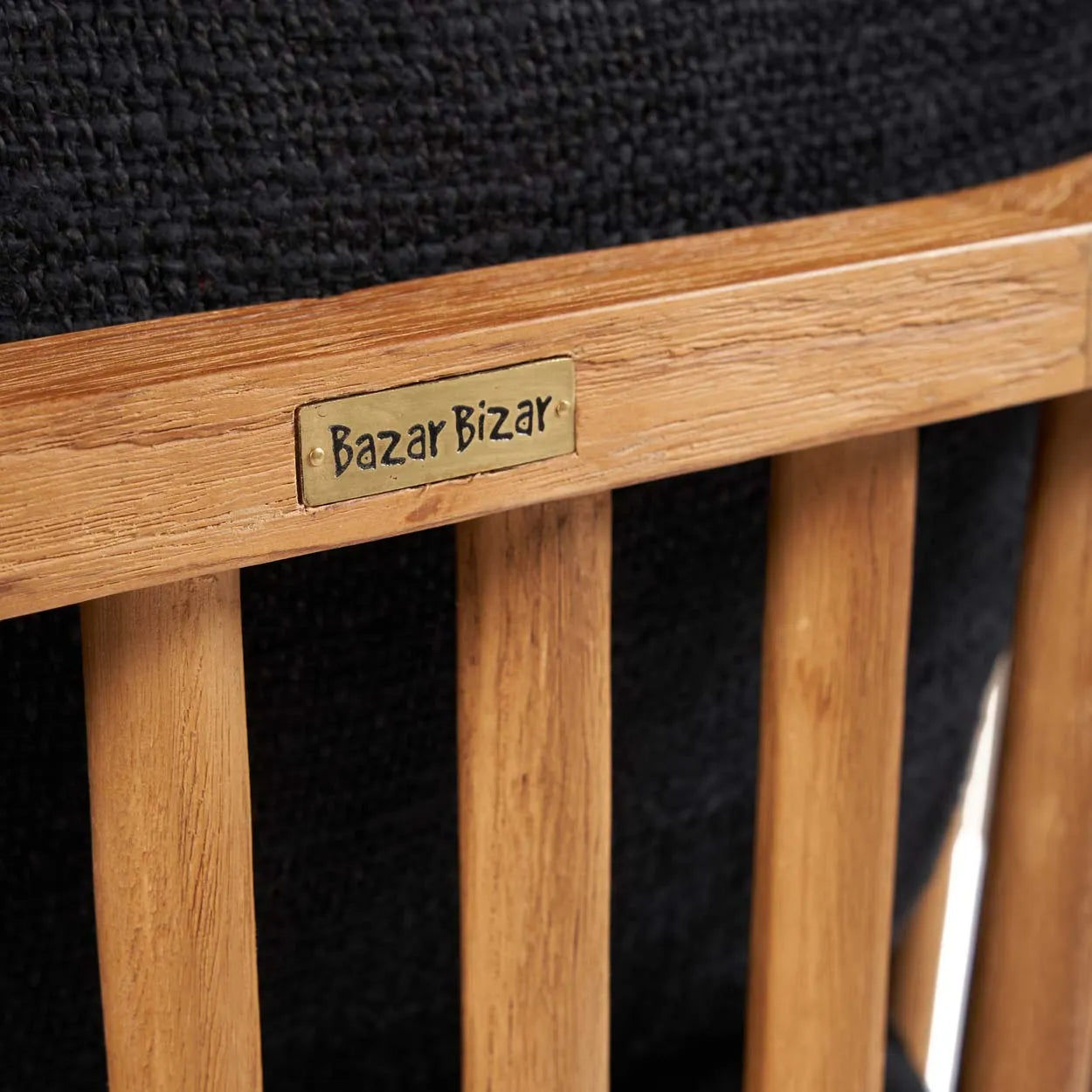 The Bazar Bizar Antwerp The Malawi One Seater - Natural Black är tillverkad av återvunnet teak med vertikala lameller. En mässingsplakett på ryggstödet har varumärket, och den är inställd mot en texturerad svart tygbakgrund, vilket förstärker dess eleganta inredningskänsla.