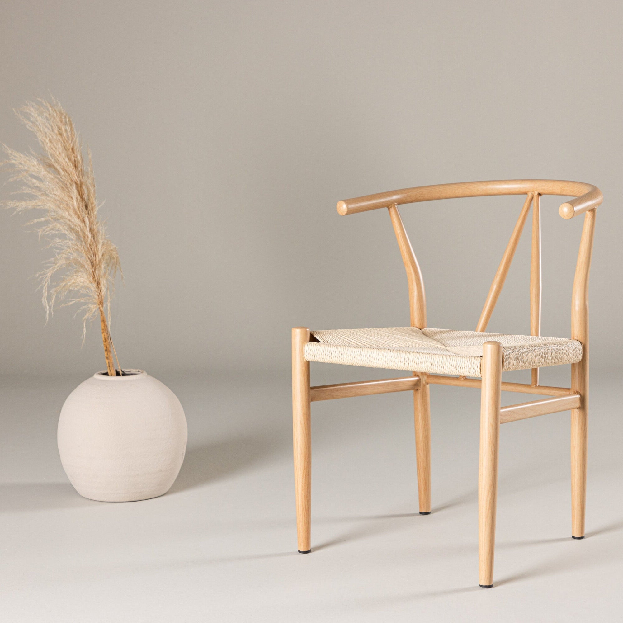 En Venture Home Alfons matstol i natur/beige rep med en sliten sits sitter på en neutral bakgrund. Till vänster lägger en rund vit vas med högt torkat pampasgräs till den minimalistiska och fridfulla moderna designen.